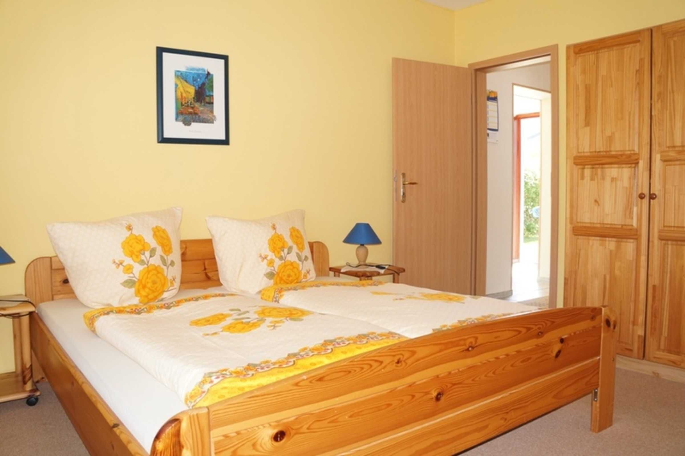 A bedroom with a wooden bed, two pillows with floral designs, a bedside table with a lamp, a framed picture on the wall, and a wardrobe.