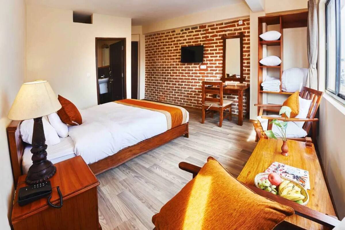 Habitación de hotel con una cama, un escritorio con lámpara, una silla, un televisor montado en una pared de ladrillo y una ventana con cortinas.
