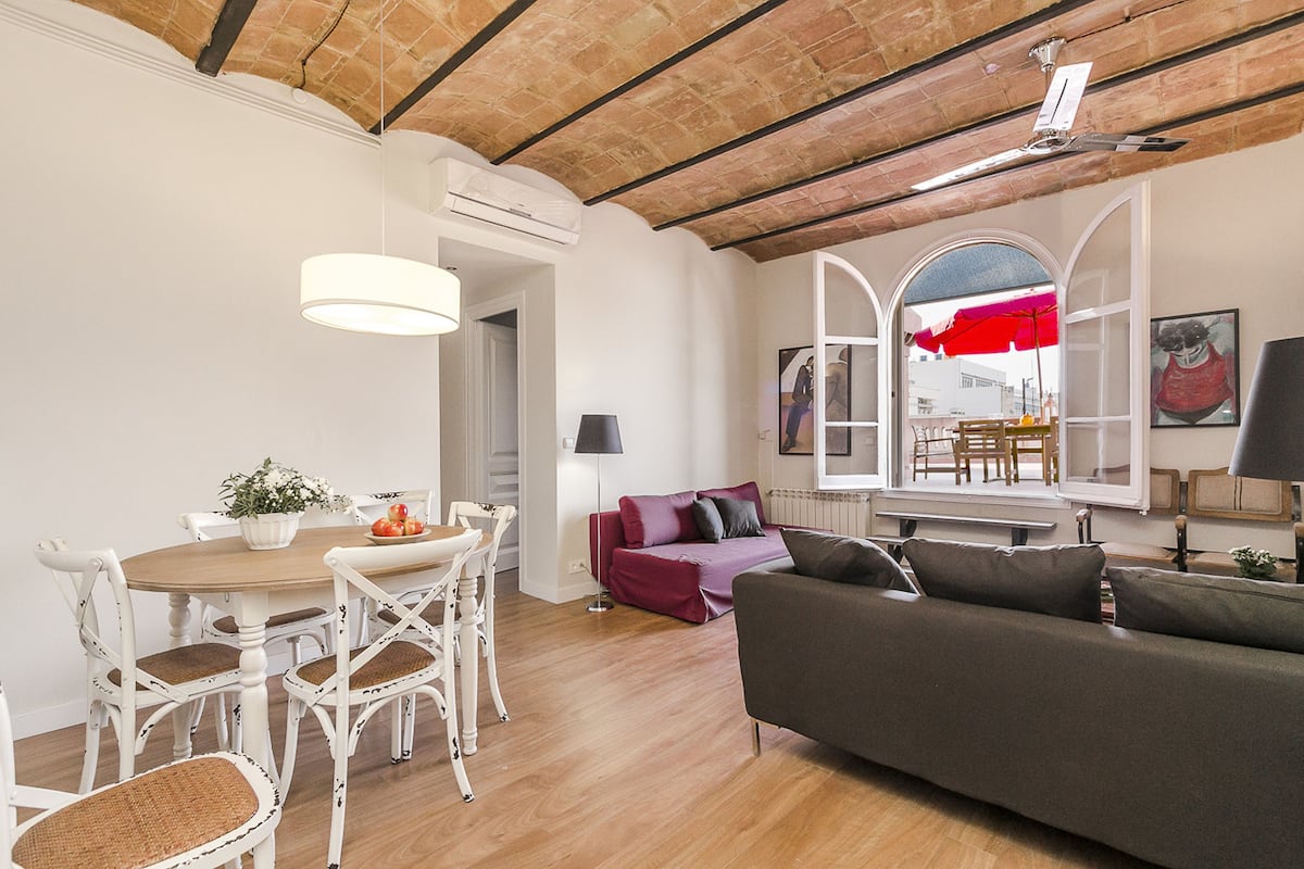 Un coin repas avec une table en bois et des chaises blanches, un canapé et, au fond, un parasol rouge.