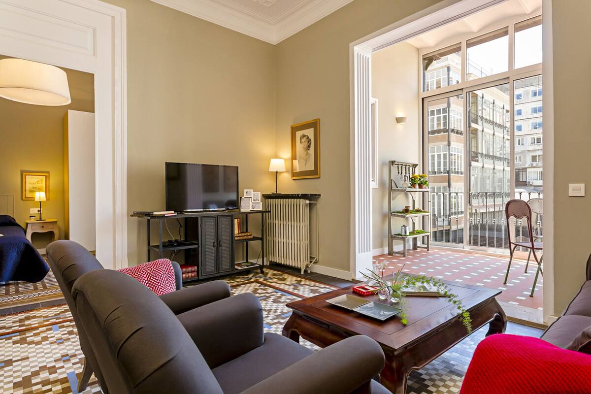 Un salon moderne avec un canapé, une table basse, un téléviseur et une grande fenêtre donnant sur des immeubles.
