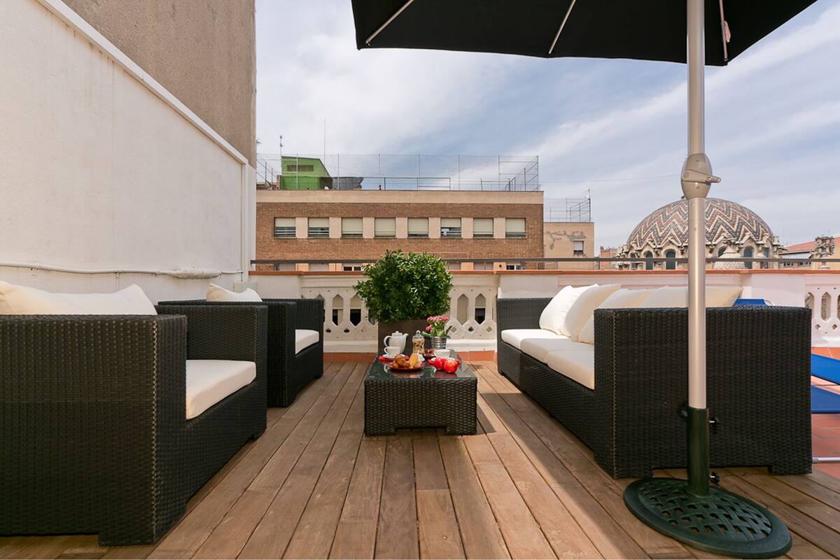 Une terrasse sur le toit, meublée de mobilier en osier, avec une table garnie de fruits et un vase, et un bâtiment en forme de dôme en arrière-plan.