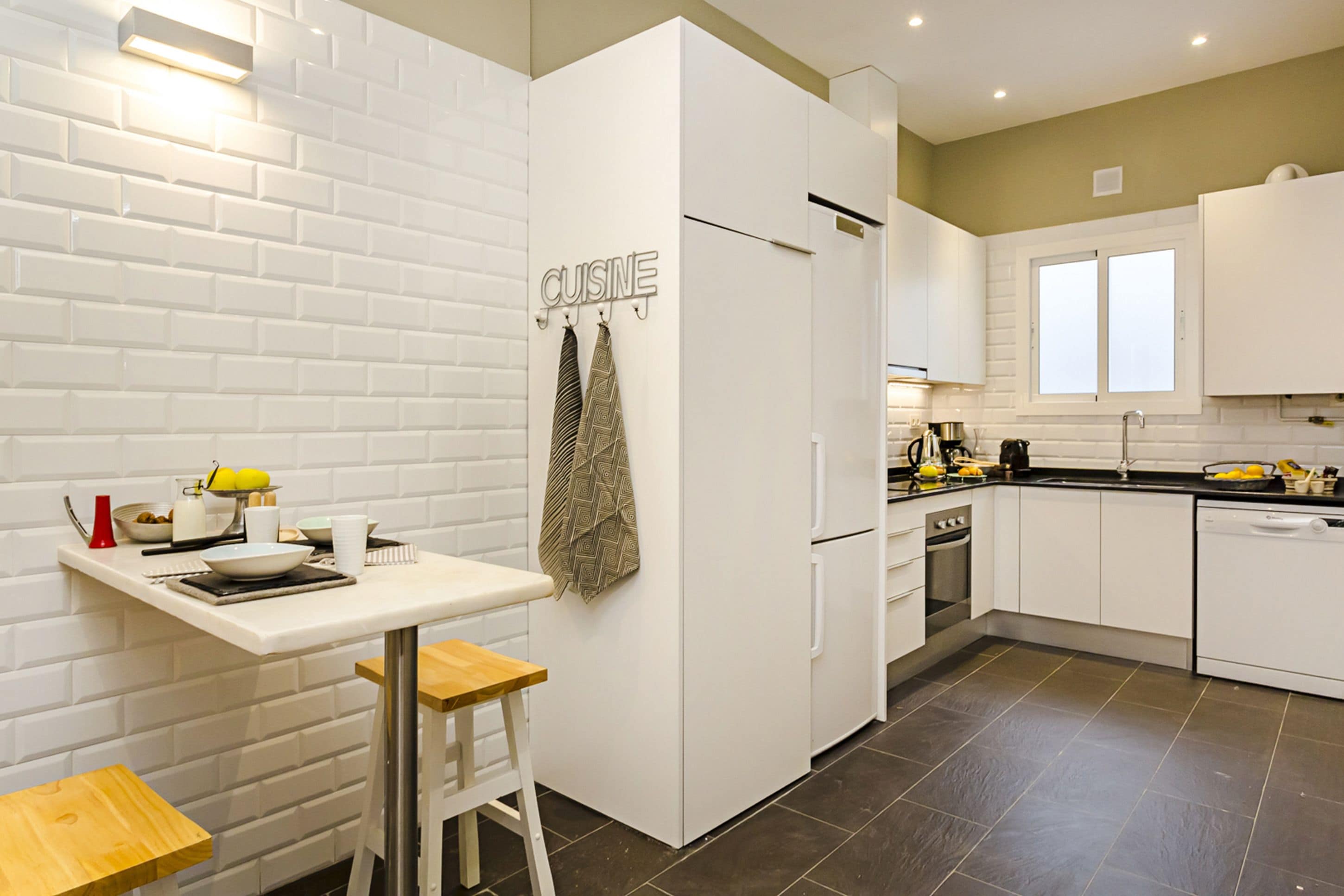 A modern kitchen with white cabinetry, a built-in oven, and a dining area with a small table and stools.