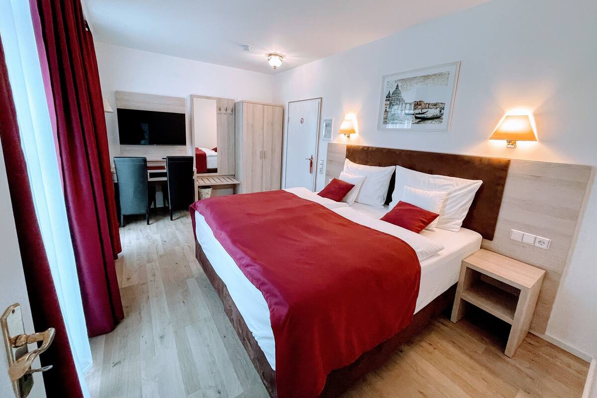 A hotel room with a large bed, a desk with a chair, a wardrobe, and a television.