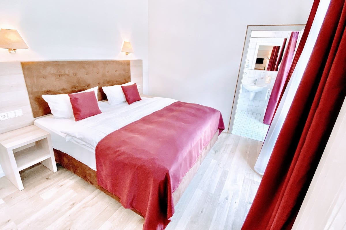 A hotel room with a large bed, white walls, a wooden headboard, and a mirror reflecting the bathroom.