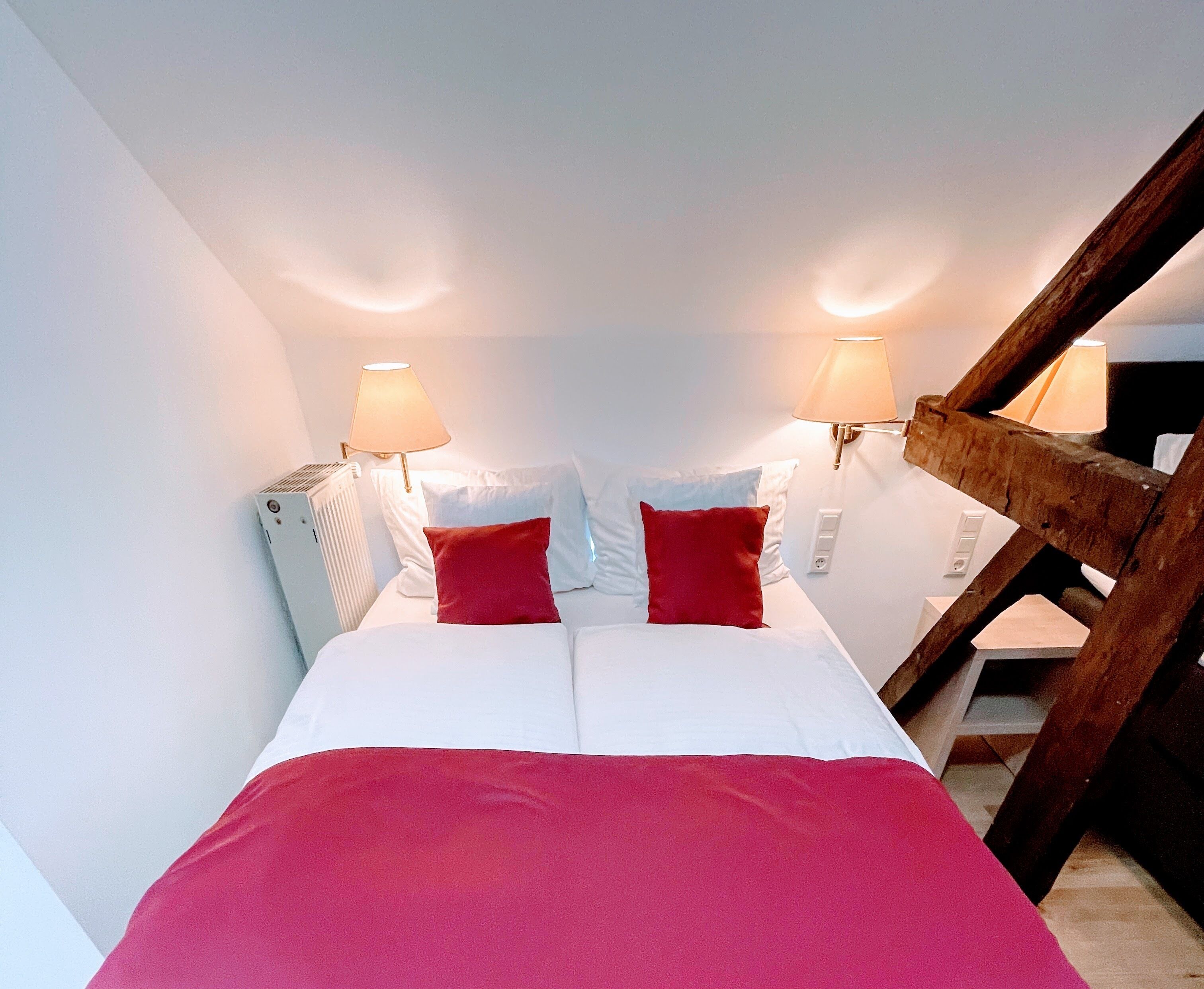 A bedroom with a bed, two red pillows, two wall-mounted lamps, and a wooden ladder.