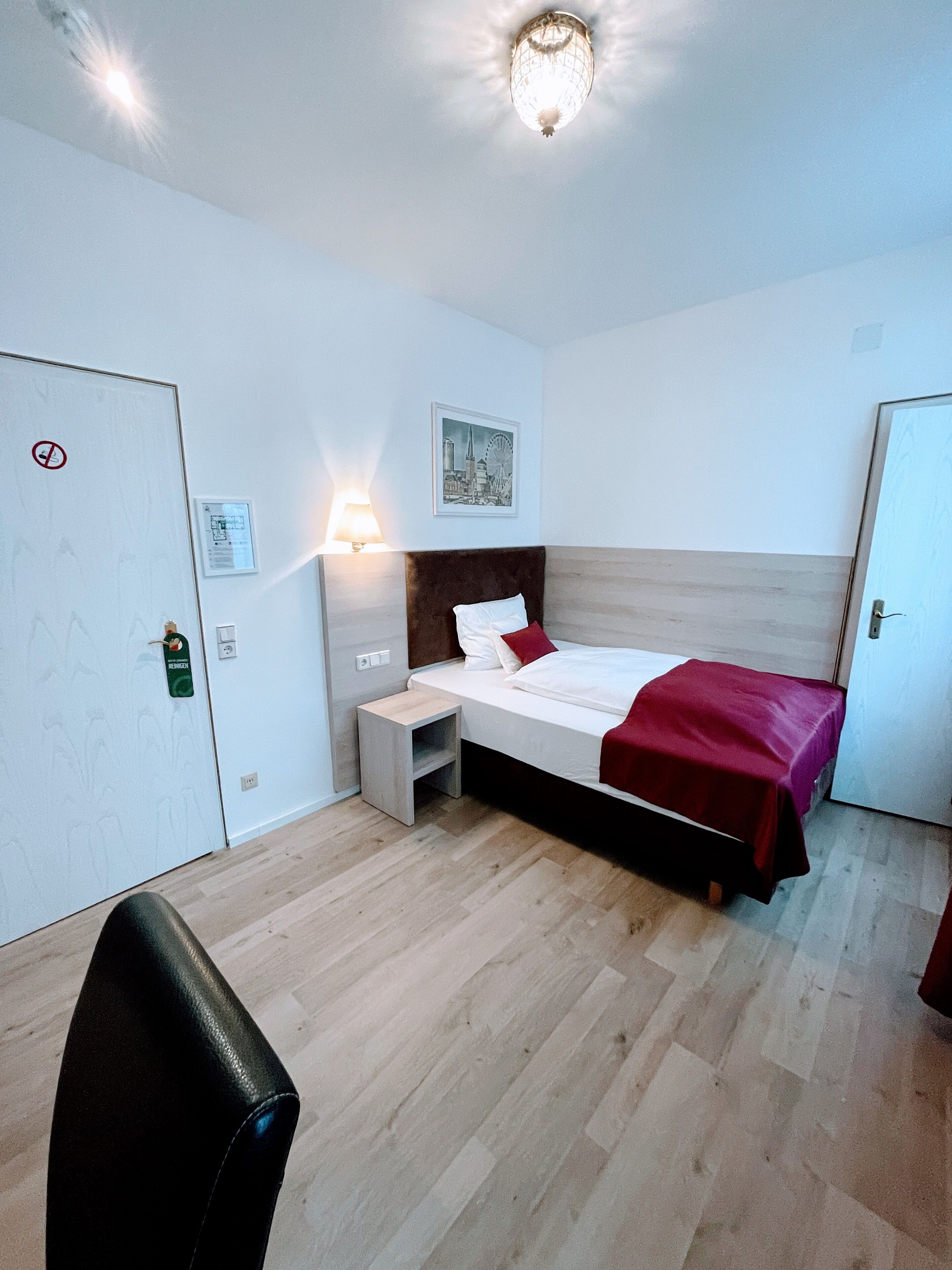 A hotel room with a bed, bedside table, and a lamp. There is a door with a no-smoking sign and a keycard holder.