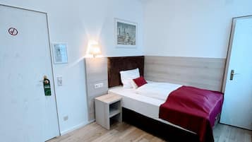 A hotel room with a bed, bedside table, and a lamp. There is a door with a no-smoking sign and a keycard holder.