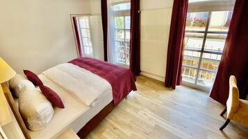 A hotel room with a bed, a wooden floor, a window with curtains, and a chair.