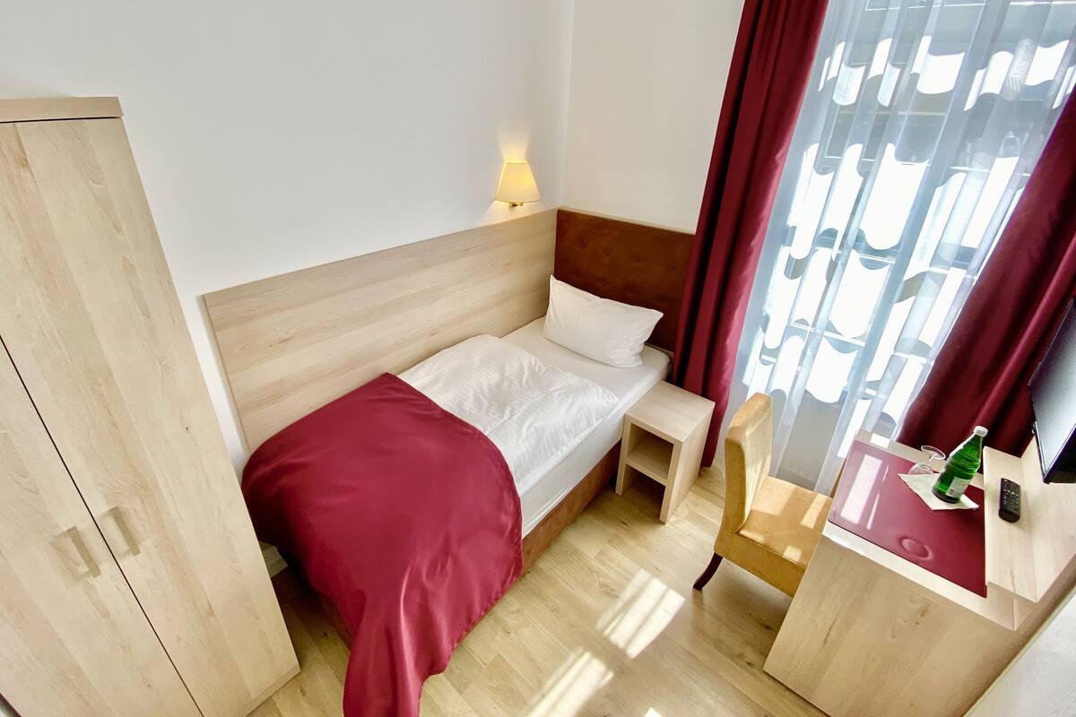 A bedroom with a wooden headboard, a bed with white bedding, a small bedside table, a chair, a desk with a computer, and a window with curtains.