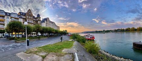 A riverside promenade with historic buildings, a red boat docked on the river, and a grassy area with people walking.