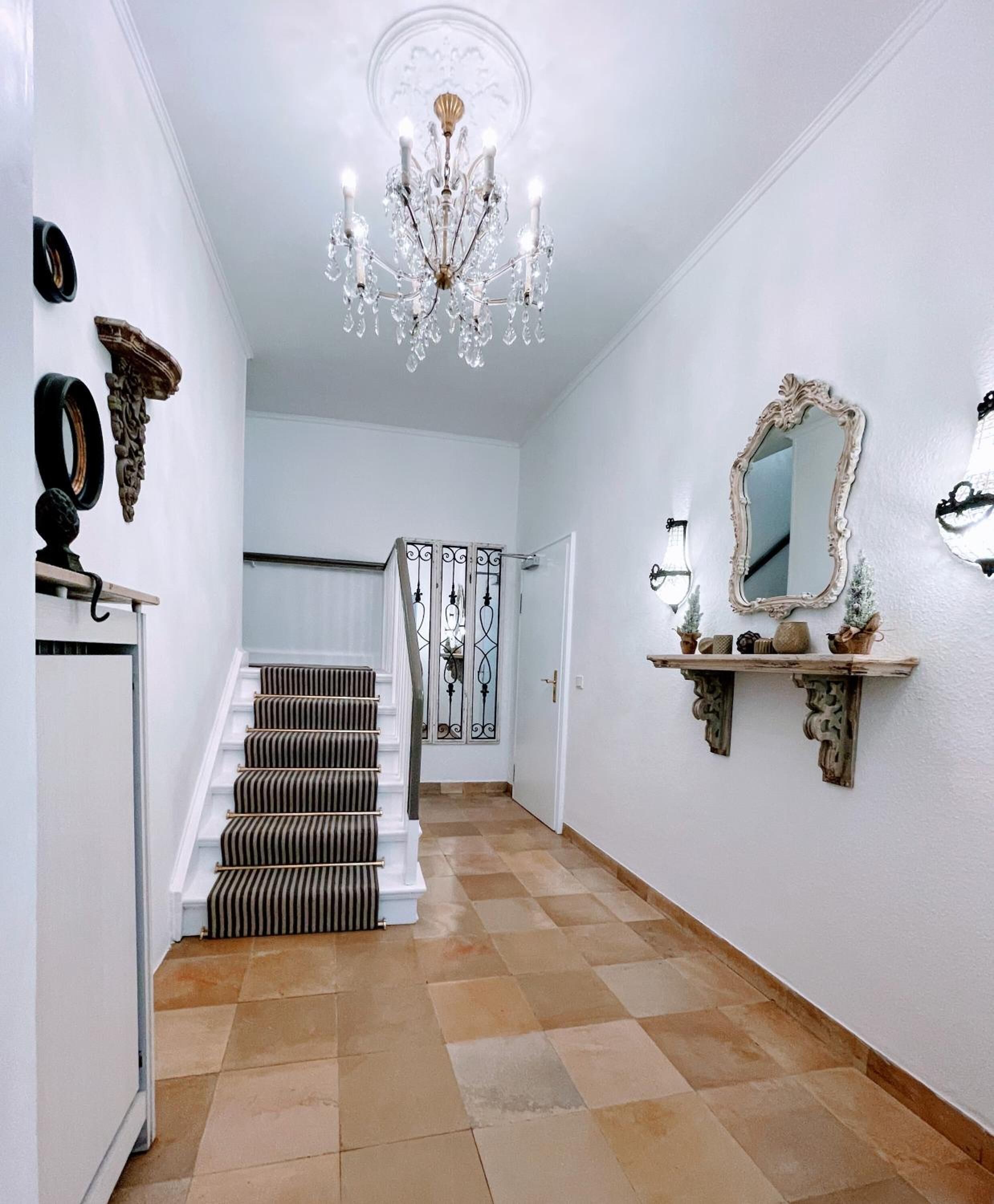 A hallway with a staircase, a chandelier, a mirror, and decorative wall elements.