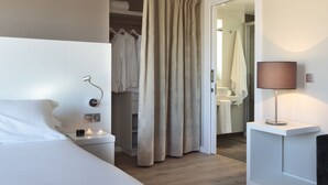 Una habitación de hotel moderna con una cama bien hecha, una lámpara de noche, un vestidor y un baño visible a través de una puerta abierta.