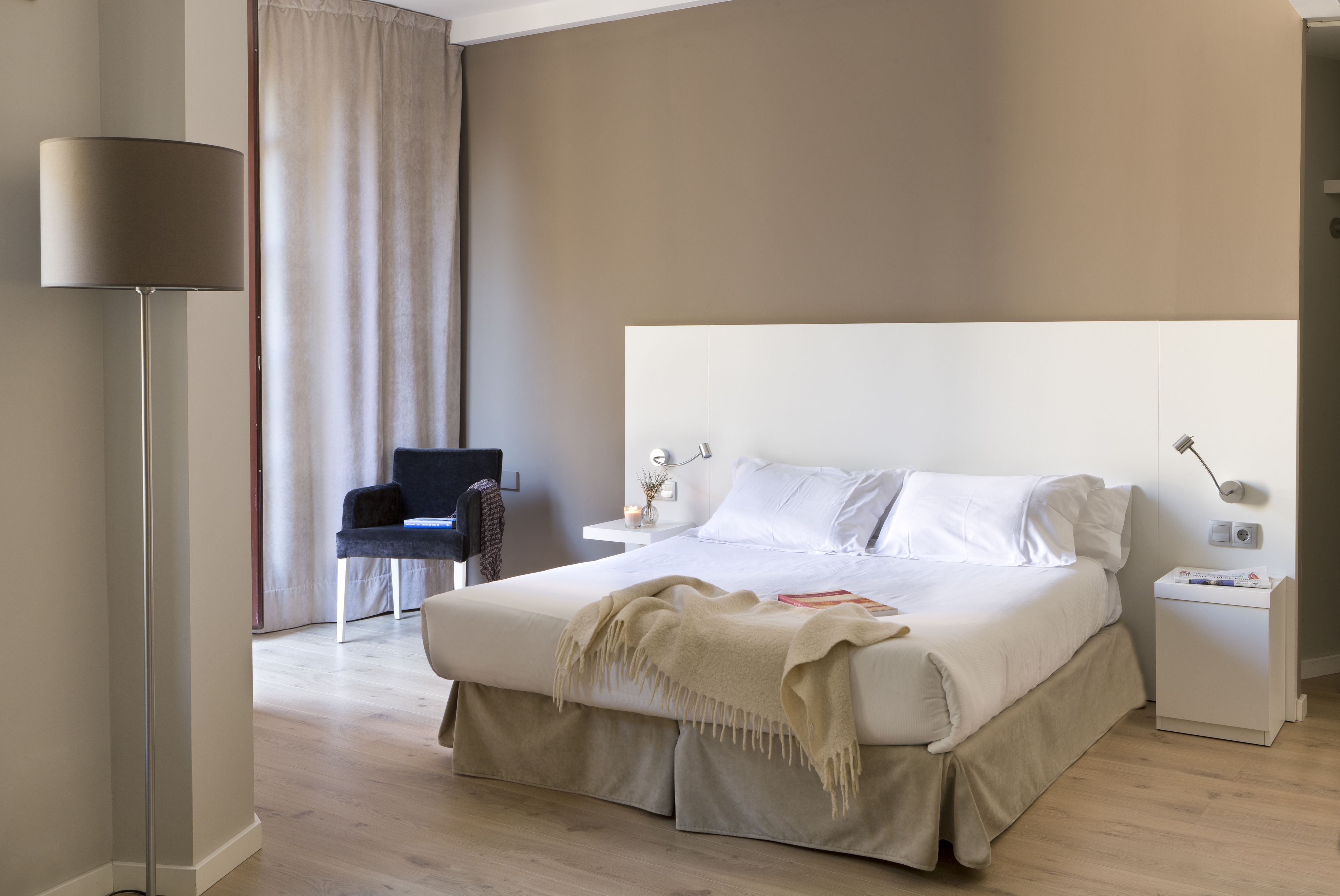 A modern hotel room with a large bed, a bedside table, a lamp, a chair, and a curtain.