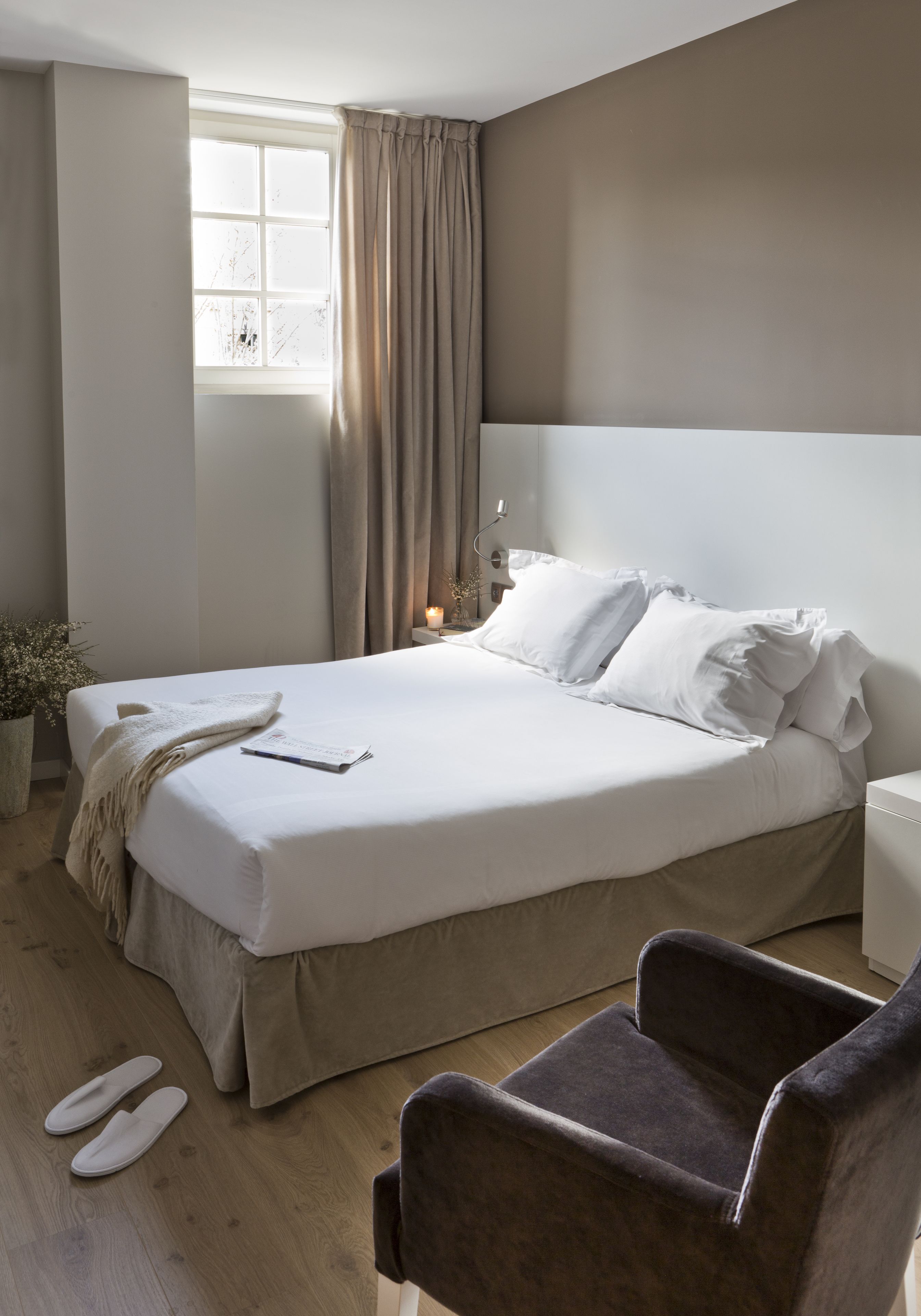 A neatly made bed with white linens, a bedside table with a lamp, and a chair in a modern room with a large window and curtains.