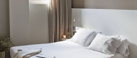 A neatly made bed with white linens, a bedside table with a lamp, and a chair in a modern room with a large window and curtains.