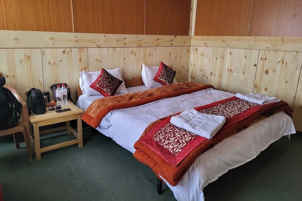 A hotel room with a wooden paneled headboard, a bed with white and red bedding, a nightstand with a bag and a water bottle, and a green carpet.