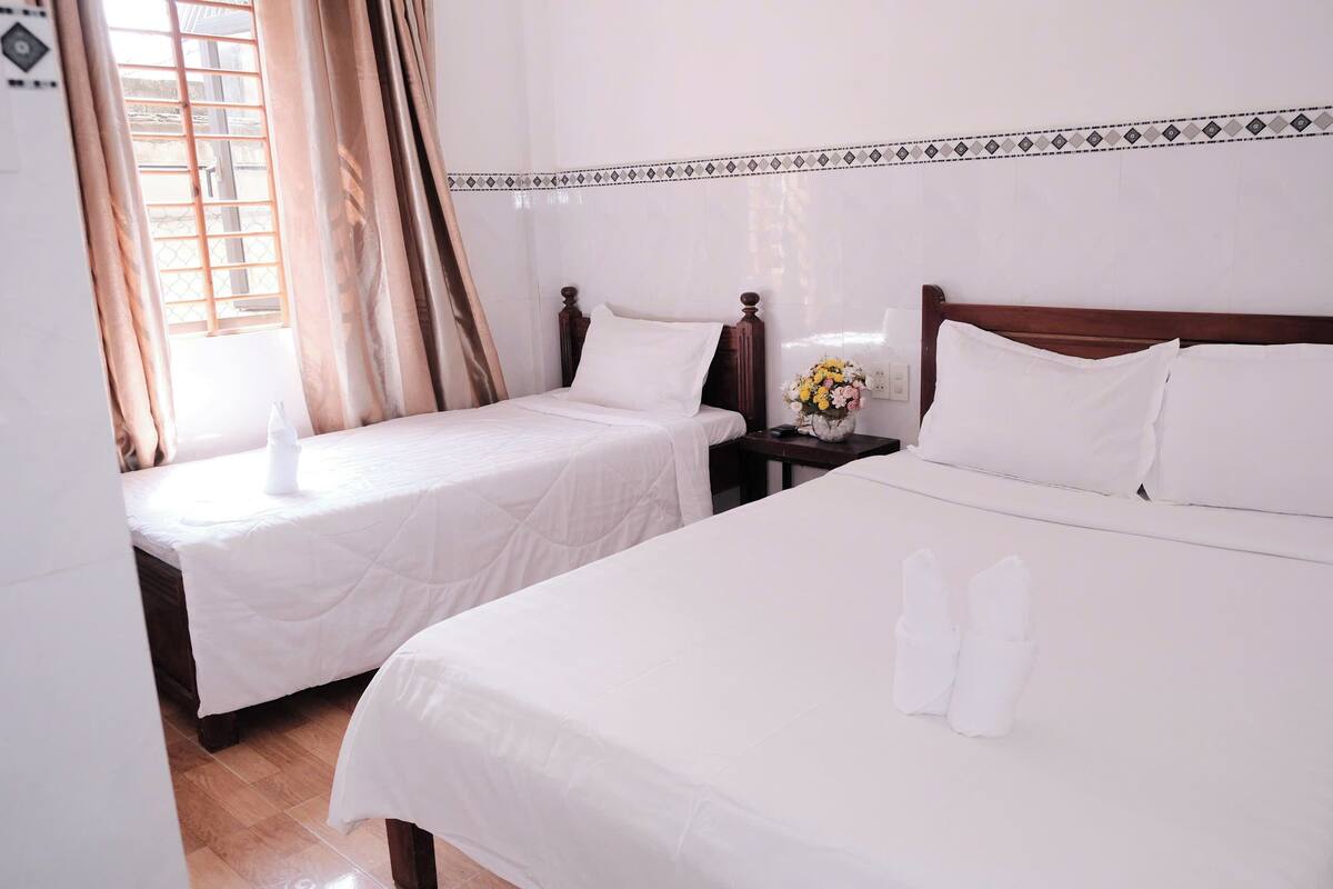 Une chambre d’hôtel avec deux lits simples, une tête de lit en bois, une table de chevet avec un vase de fleurs et une fenêtre avec des rideaux.
