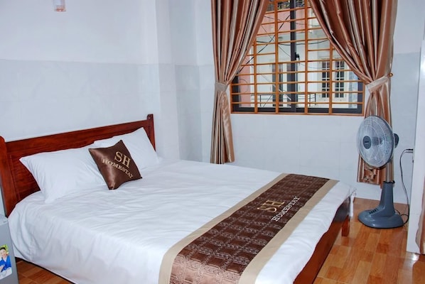A neatly made bed with a brown pillow, a bedside fan, and a window with curtains.