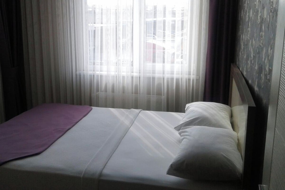 Una cama individual con colchón blanco, manta morada, almohada blanca y cortinas translúcidas que dejan pasar la luz natural.