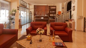 A well-lit living room with red sofas, a coffee table with decorative items, and a reception desk with a wooden partition. - Udi Hotel (Çanakkale)