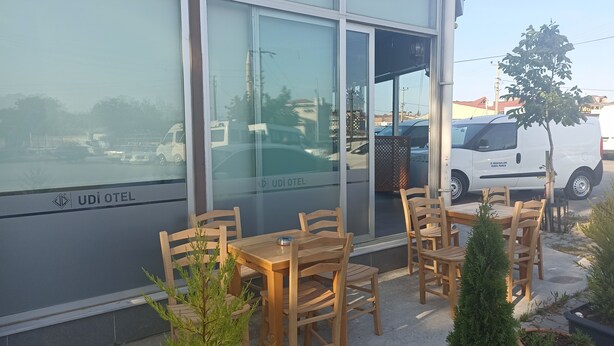 An outdoor seating area with wooden tables and chairs in front of a building with large windows. - Udi Hotel (Çanakkale)