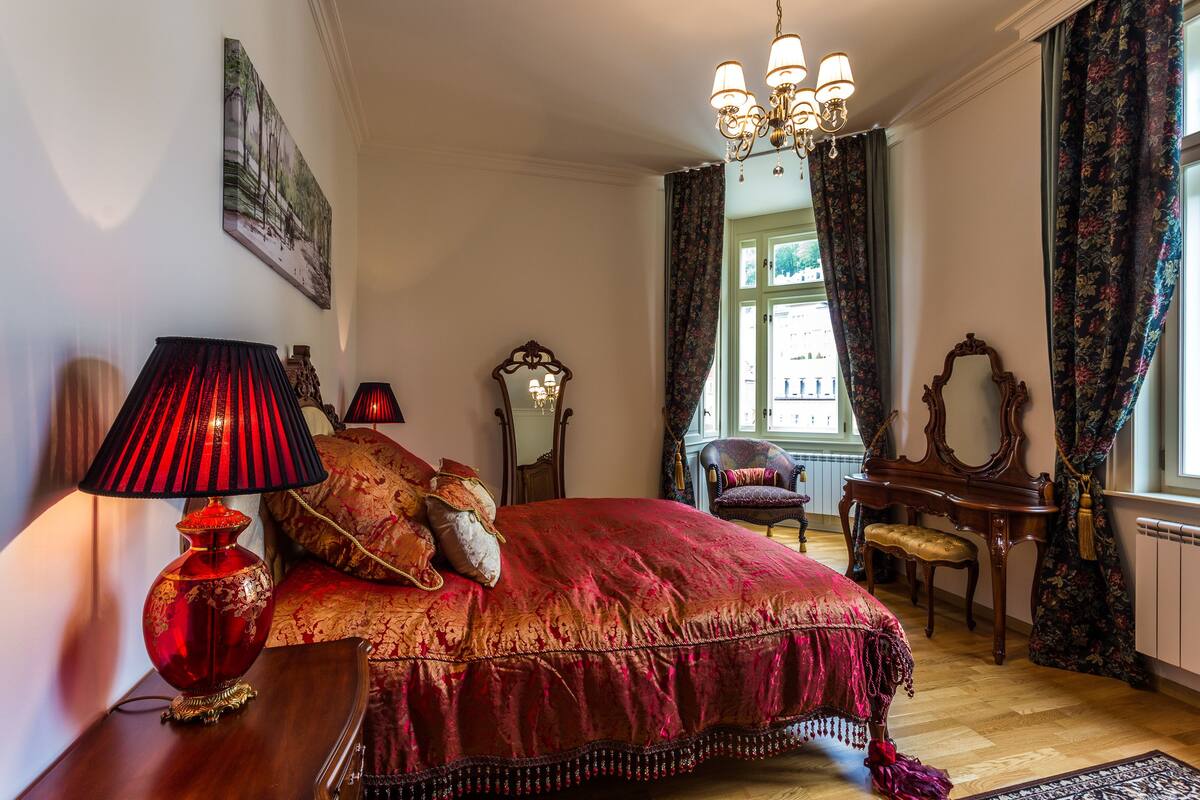A bedroom with a large bed, a wooden dresser, a chair, a piano, and a window with curtains.