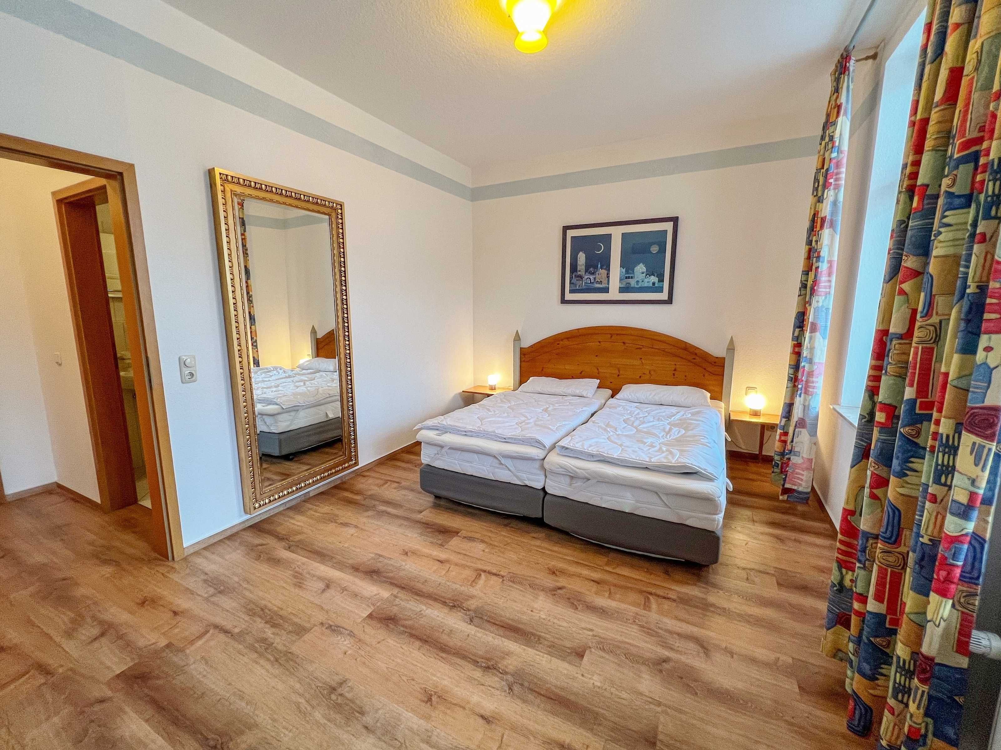 Habitació d'hotel amb terra de fusta, un llit de matrimoni amb capçalera, un mirall gran i un quadre a la paret.