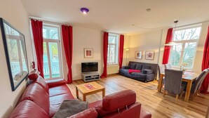 A living room with a red sofa, a TV, a grey sofa, and a dining area with a table and chairs. - Kurhaus Nordstrand - Apartment 45407 - KH - Apartment 38 (Göhren)