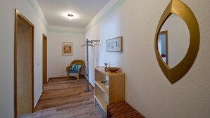 A hallway with wooden flooring, a wicker chair, a floor lamp, a shelf with a bowl, and a wall-mounted mirror. - Kurhaus Nordstrand - Apartment 45407 - KH - Apartment 38 (Göhren)