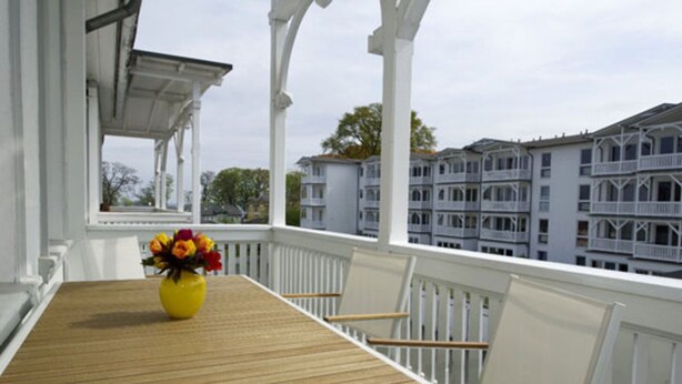A wooden table with a yellow vase of flowers on a balcony with white railings and white chairs. - Kurhaus Nordstrand - Apartment 45407 - KH - Apartment 38 (Göhren)