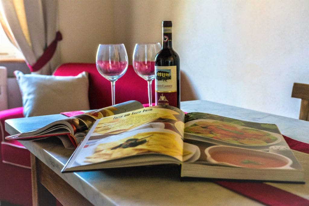 Une table avec une bouteille de vin et deux verres, un magazine ouvert à une page consacrée à la gastronomie, et un canapé rouge en arrière-plan.