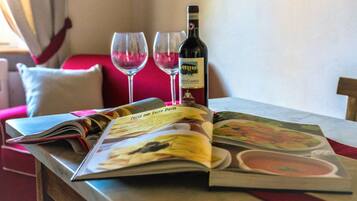 Un tavolo con una bottiglia di vino e due bicchieri, una rivista aperta su una pagina di ricette, e un divano rosso sullo sfondo.