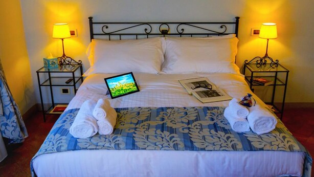 A neatly made bed with a tablet, two lamps, and towels on a floral-patterned bedspread. - Palazzo Mari (San Gimignano)