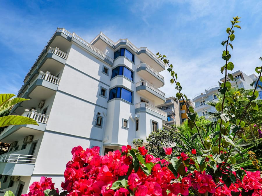 Un hotel de diverses plantes amb balcons, envoltat de vegetació exuberant i flors colorades.