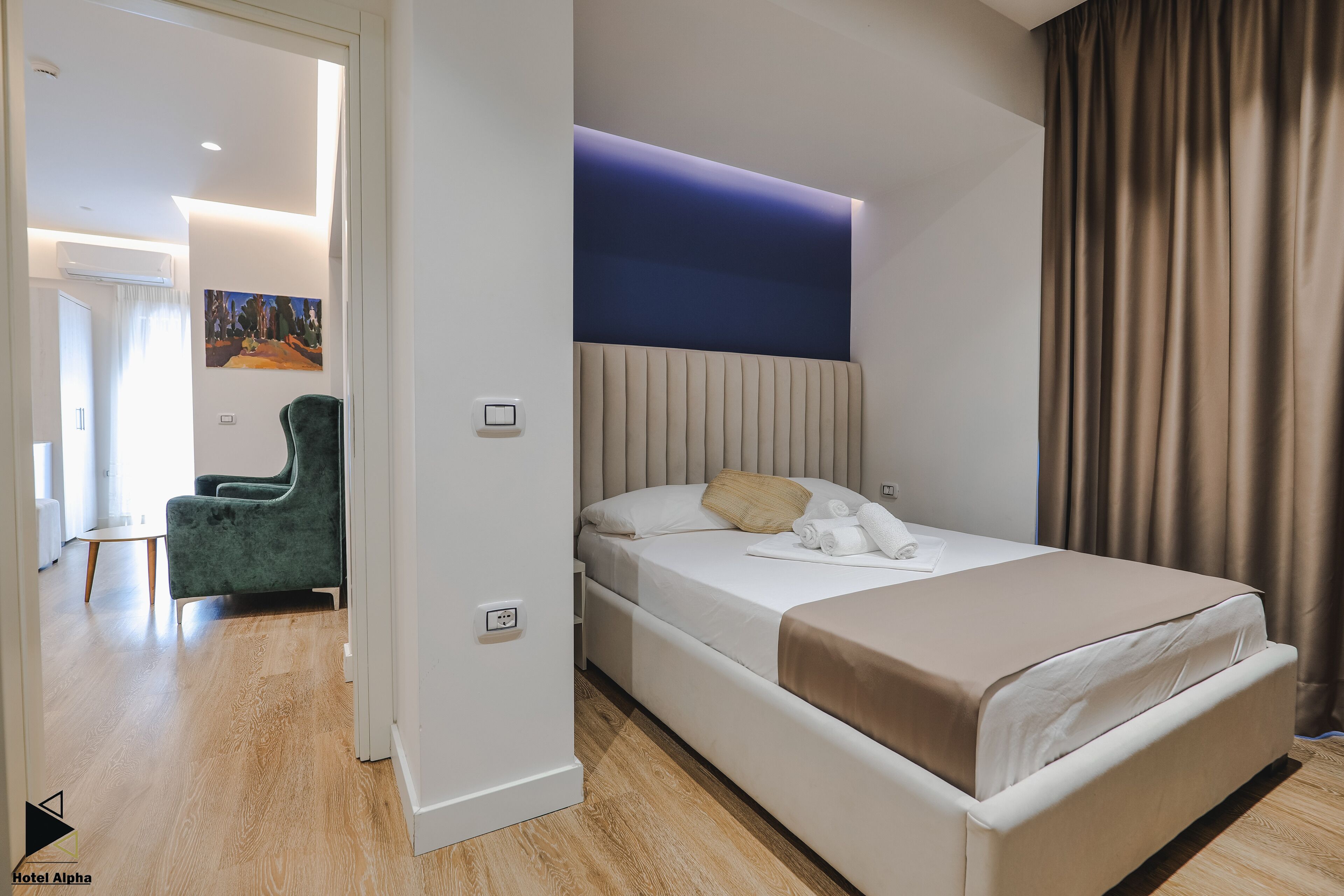 A modern hotel room with a bed, a green armchair, and a painting on the wall.