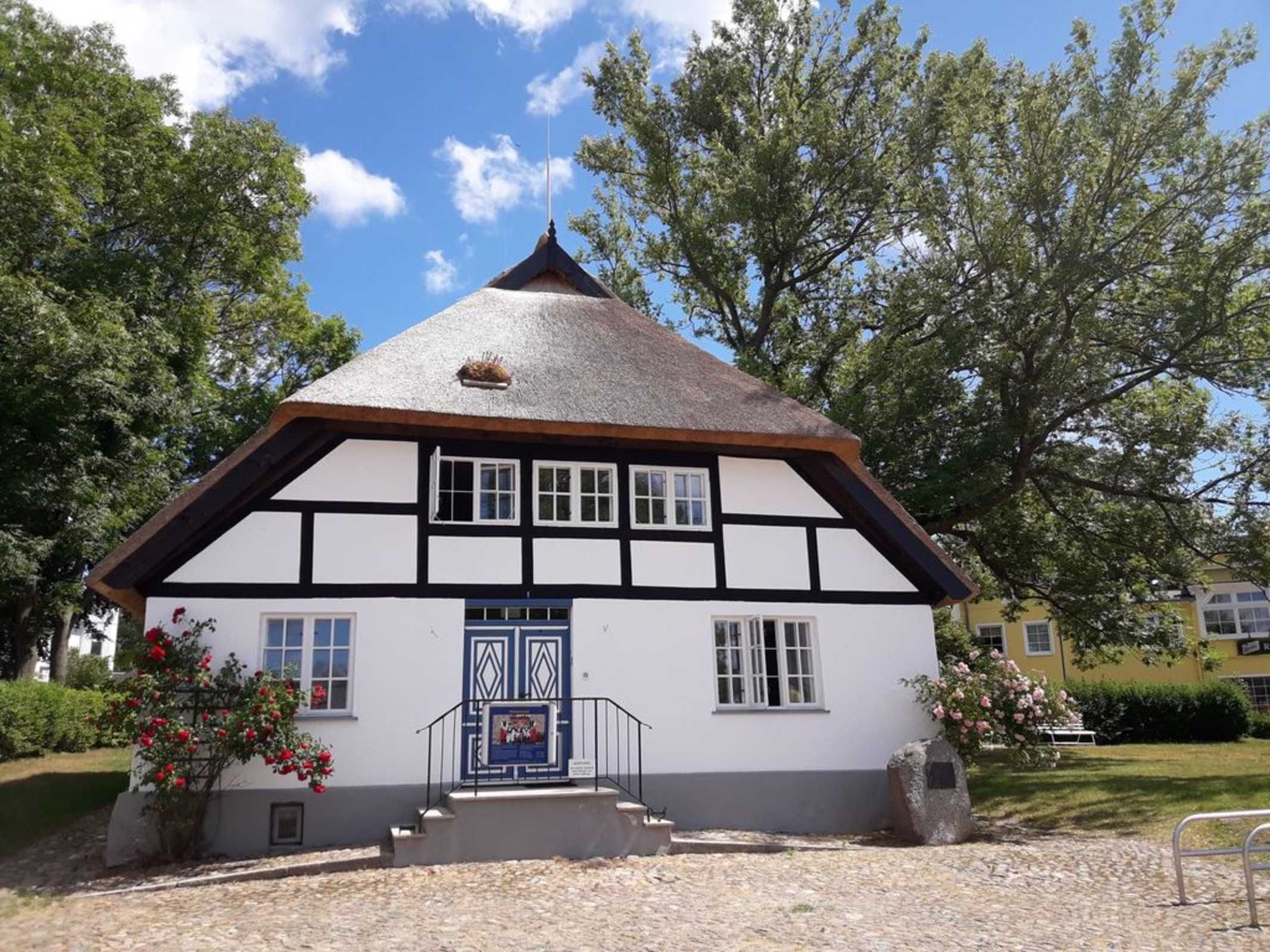 Ett traditionellt hus med korsvirkesfasad och ett brant tak, en central entré med en blå dörr och en liten veranda.