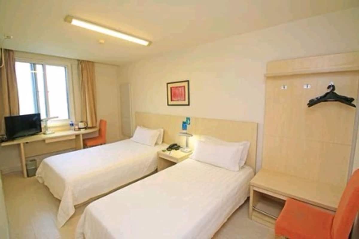 A hotel room with two single beds, a desk with a computer, a chair, a bedside table with a lamp, and a wardrobe with a hanging rack.