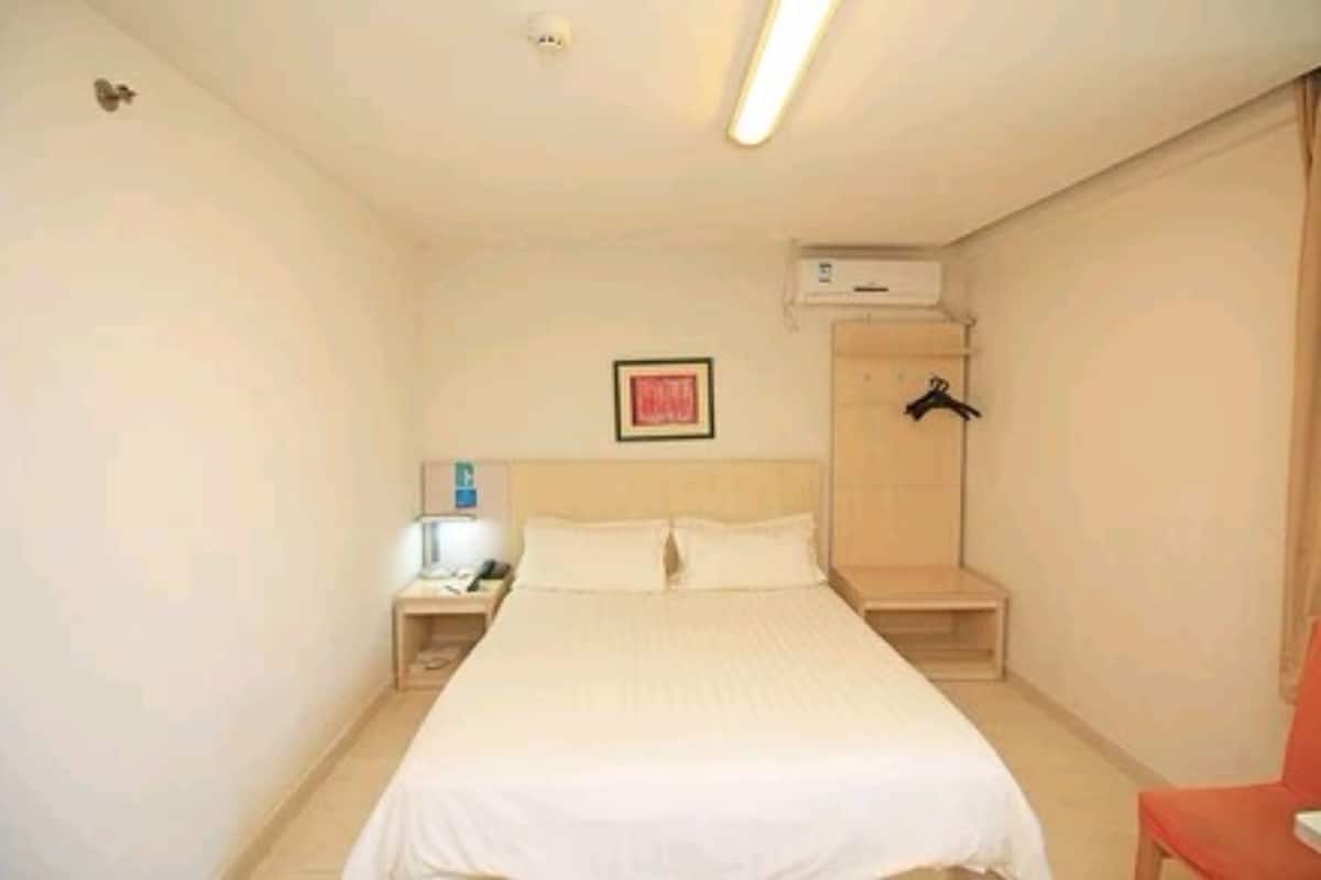 A small, clean hotel room with a single bed, a nightstand, a red chair, and a wall-mounted air conditioning unit.
