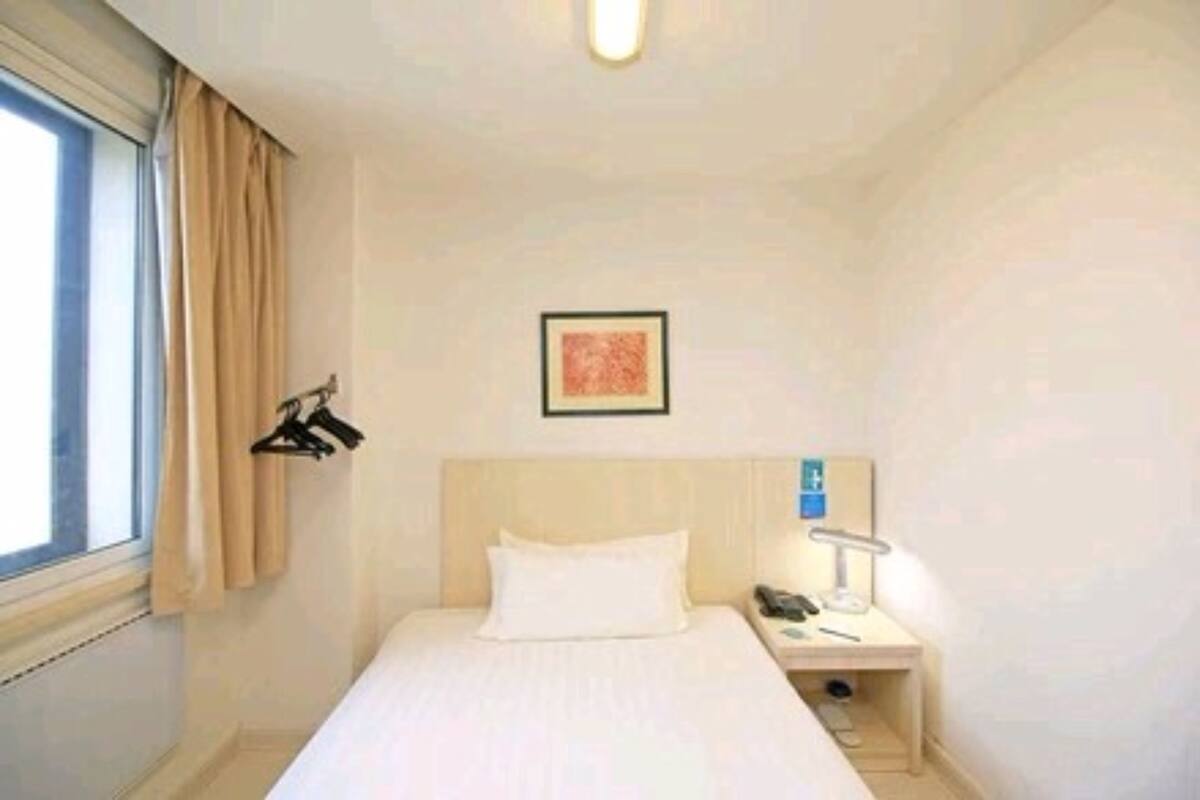 A hotel room with a bed, a bedside table with a lamp, a framed picture on the wall, and a window with curtains.