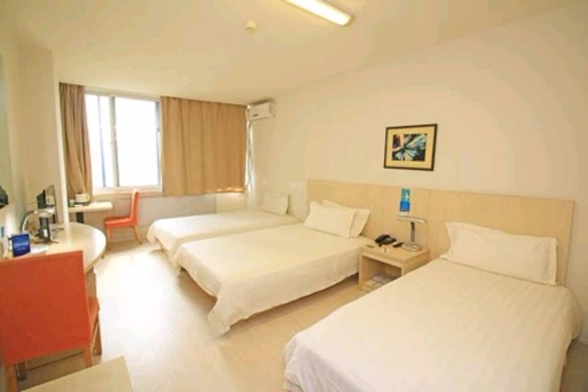 A hotel room with two beds, a desk, a chair, a window with curtains, a bedside table with a lamp, and a framed picture on the wall.