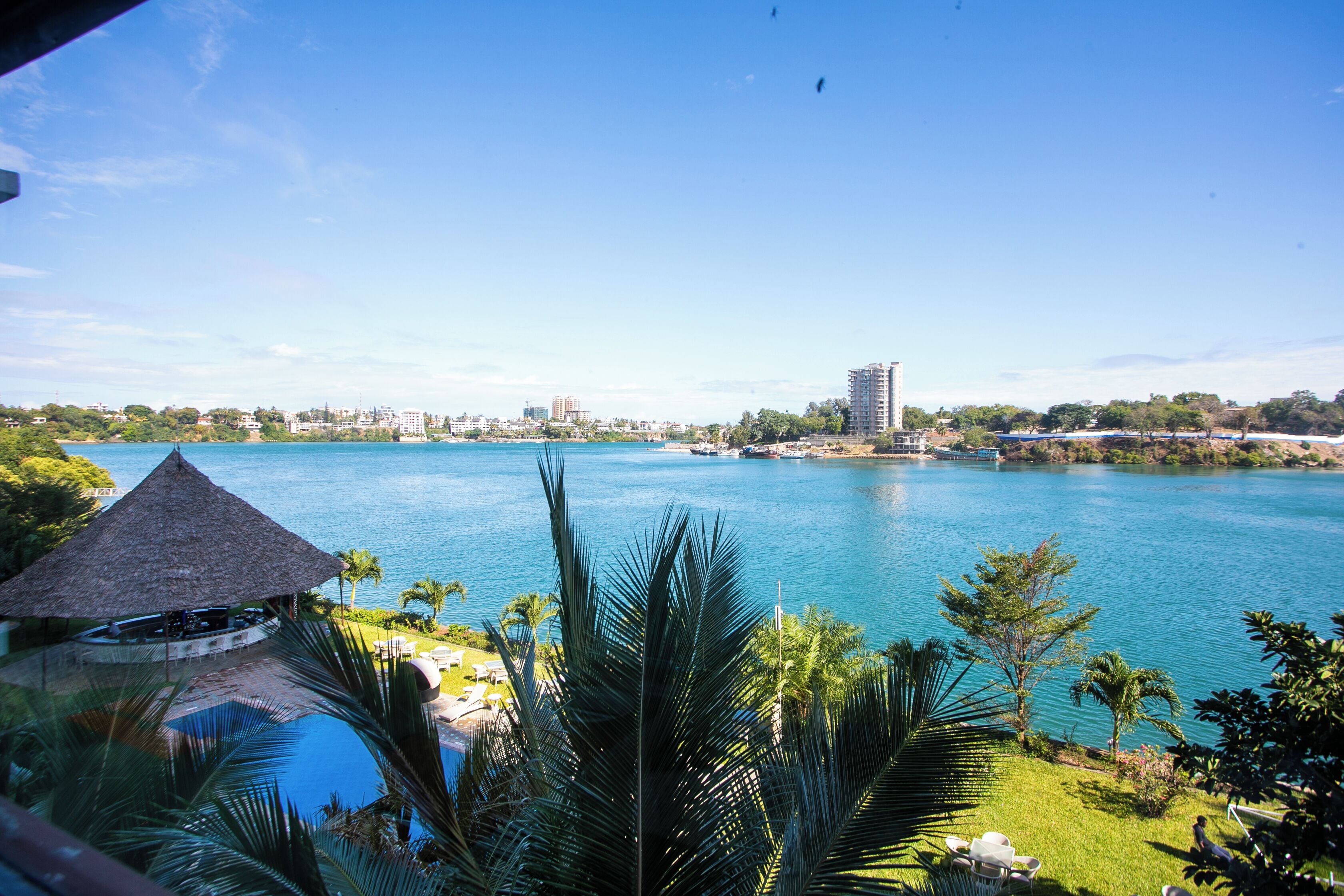 A view of a waterfront property with a pool, lounge chairs, and a thatched-roof structure.