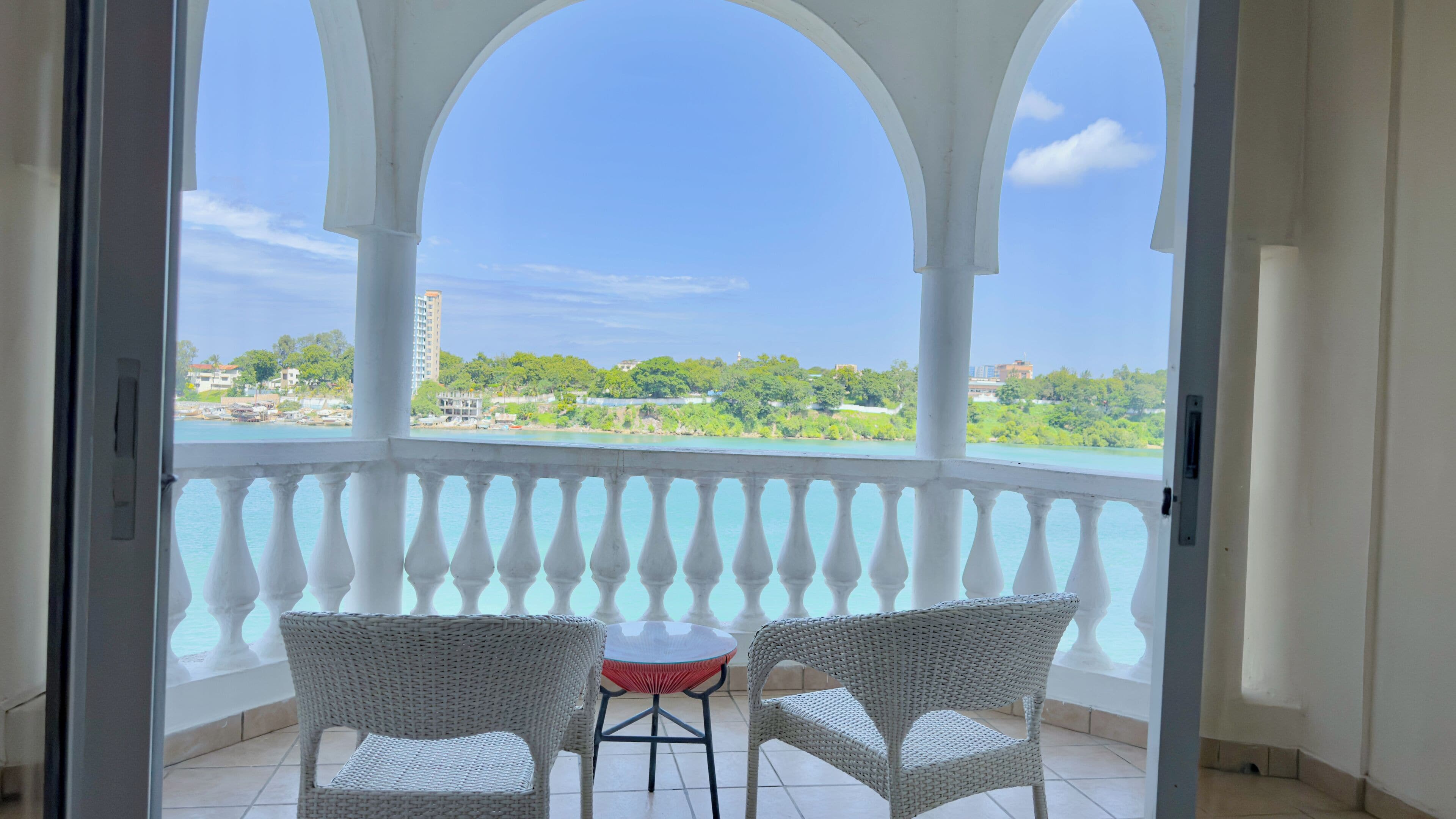 A balcony with a view of a body of water and buildings, two wicker chairs, and a small round table.