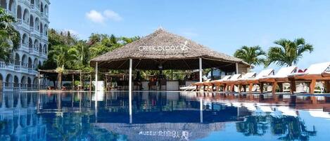 Ein Hotelpoolbereich mit einem strohgedeckten Pavillon, Liegestühlen und einem Reflexionsbecken.