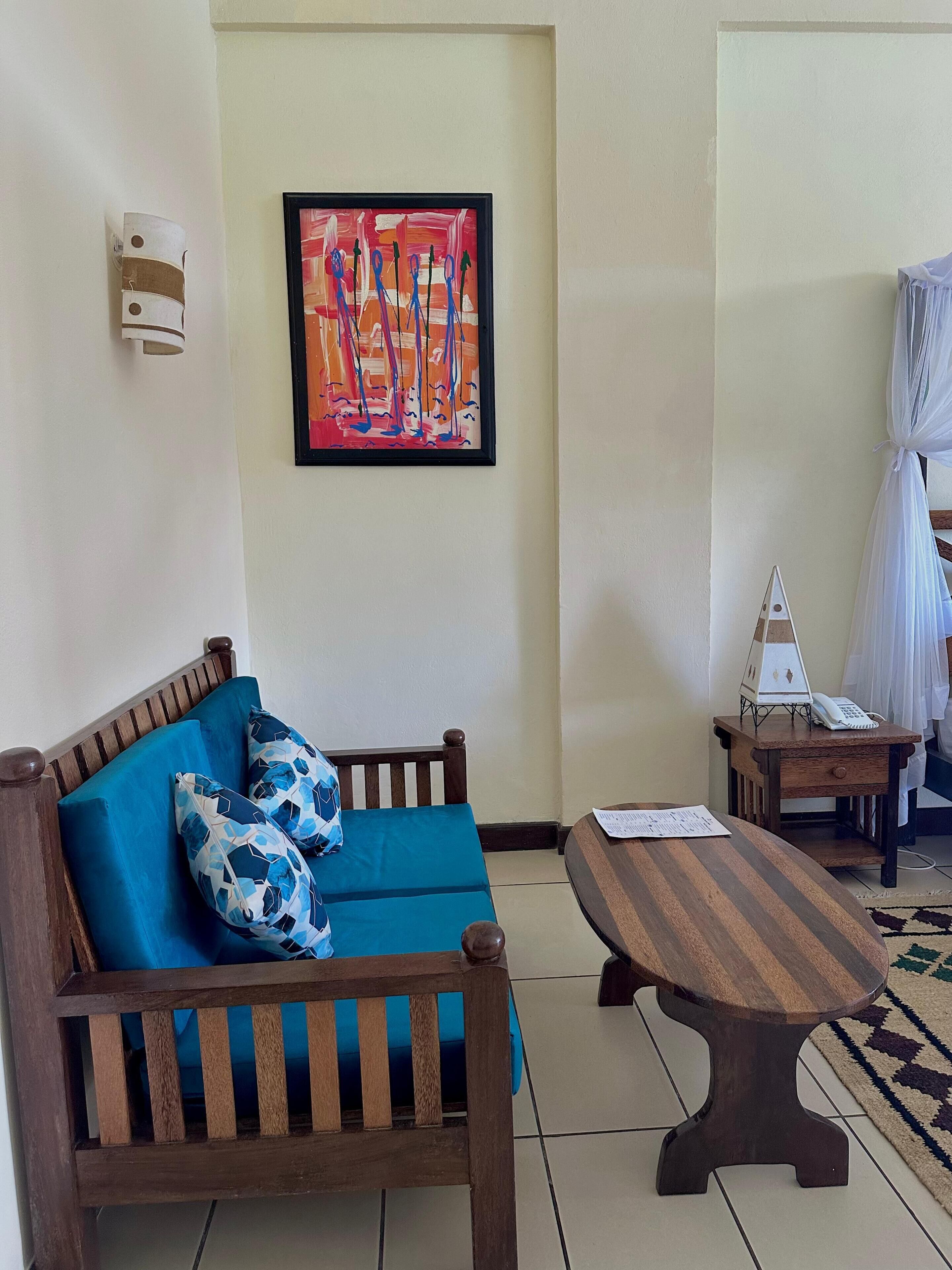 Une pièce comprenant un canapé en bois, une petite table en bois, un tableau abstrait encadré, un luminaire fixé au mur et un petit objet décoratif posé sur une table de chevet en bois.