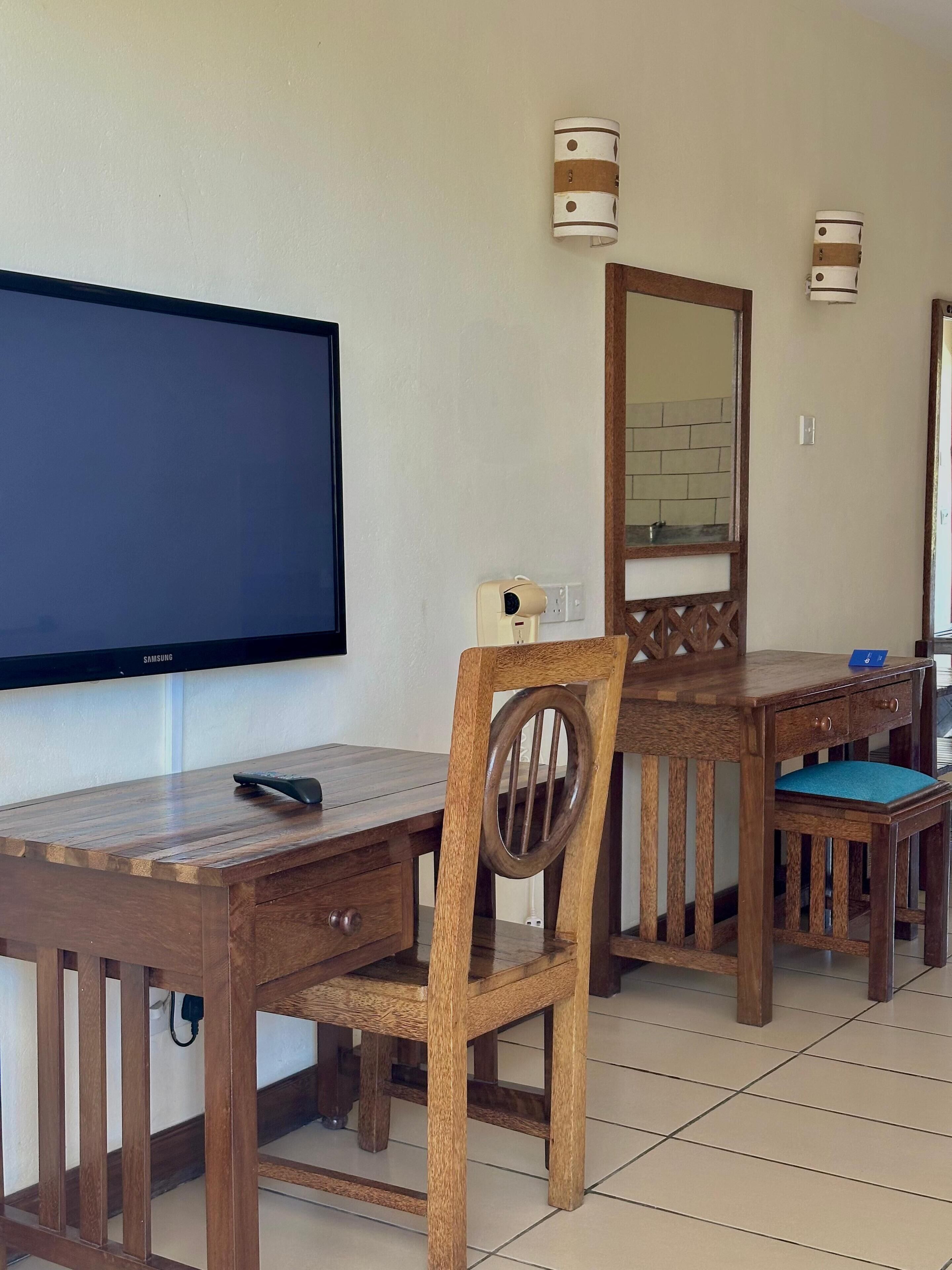 Quarto de hotel com escrivaninha de madeira, televisão, espelho e luminária na parede.