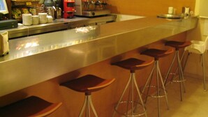 A bar area with a stainless steel counter, stacked plates, a coffee machine, and bar stools. - Hotel Estel (Berga)