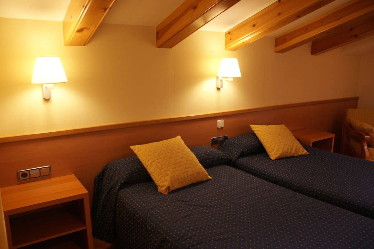 A hotel room with two beds, each with a pillow and a lamp on the wall. There is a wooden headboard, a nightstand, and a chair.