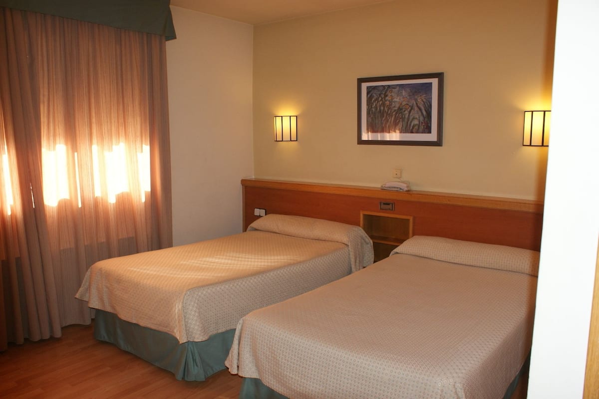 A hotel room with two beds, a painting on the wall, and two wall-mounted lamps.