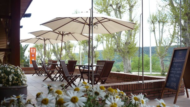 An outdoor seating area with tables and chairs under umbrellas, a signboard with menu options, and a view of trees and hills. - Hotel Estel (Berga)