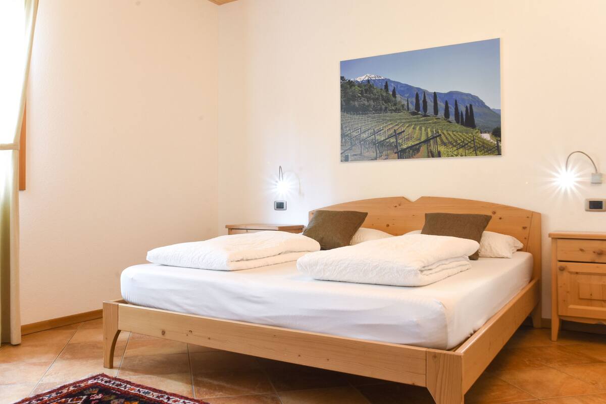A bedroom with a wooden bed, white linens, a nightstand, a wall-mounted lamp, and a painting of a vineyard.